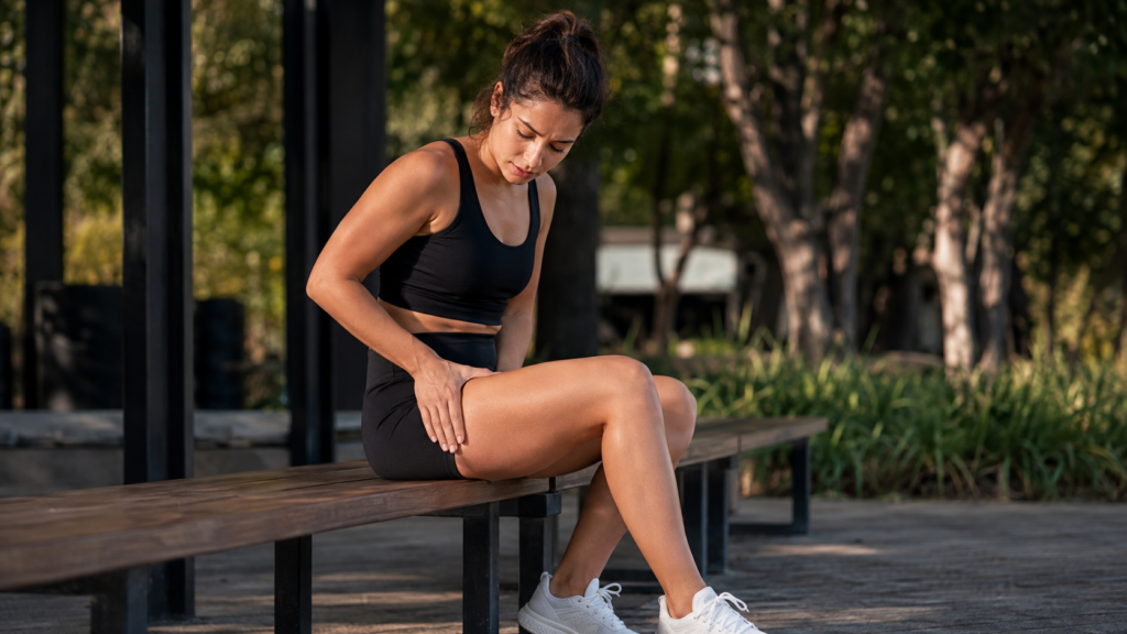 Dor na frente da coxa depois do treino: o que pode ser? Mulher após o treino tocando a parte da frente da coxa devido a desconforto muscular.